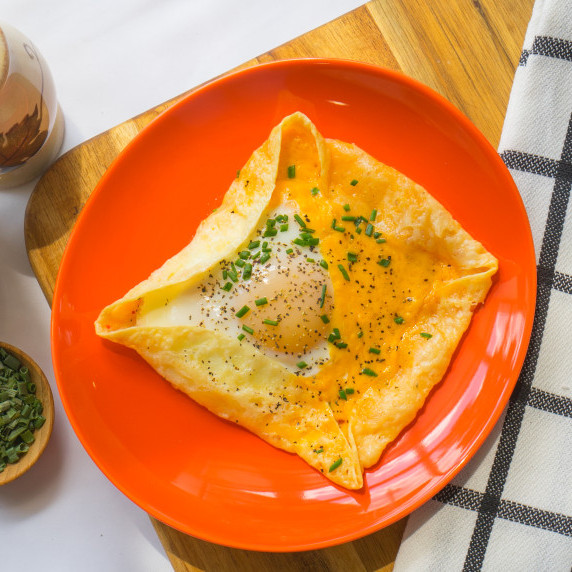 Egg galette in a cheese wrap, topped with chives, on red plate on top of a cutting board