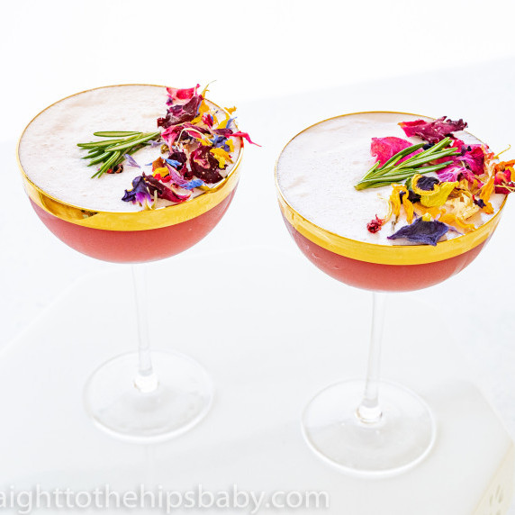 two cocktail coupes filled with a red cocktail with white foam. Topped with dried flower petals