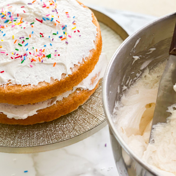 A cake in the back covered in frosting and a bowl with frosting in the front. 