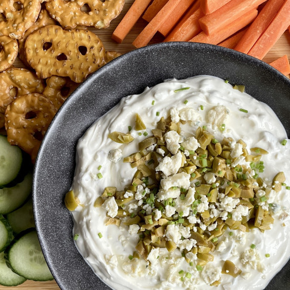 Gray bowl filled with a creamy olive and blue cheese dip next to pretzels, carrots, and cucumbers. 
