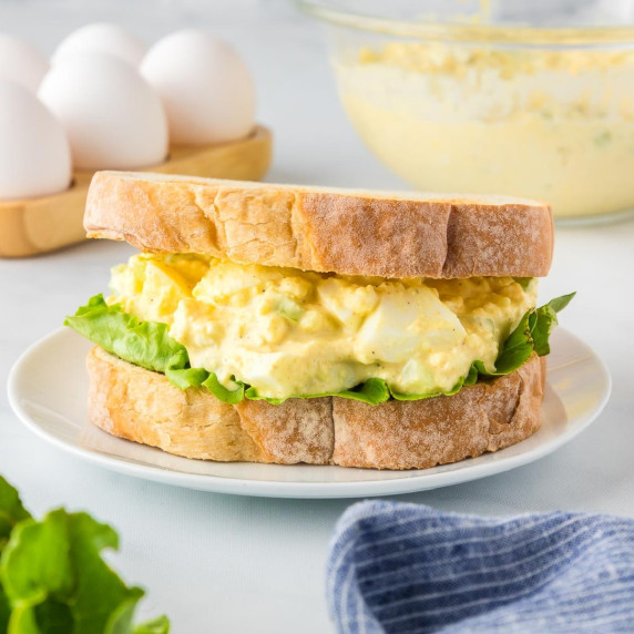A sandwich with egg salad and lettuce on thick, toasted bread is on a white plate.