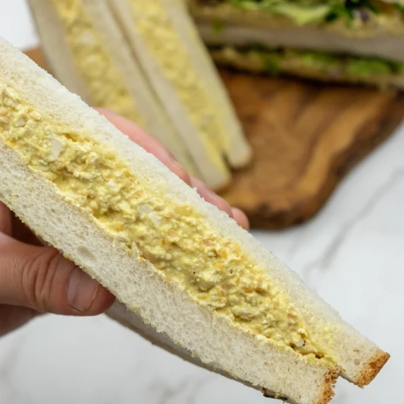 Hand holding a vegan egg salad sandwich, with more sandwich halves in the background