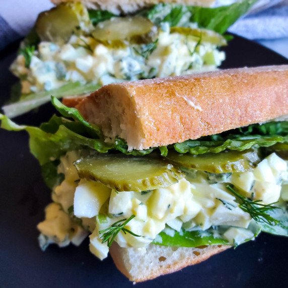 Creamy egg salad and crisp, green veg on a toasted baguette against a black background.
