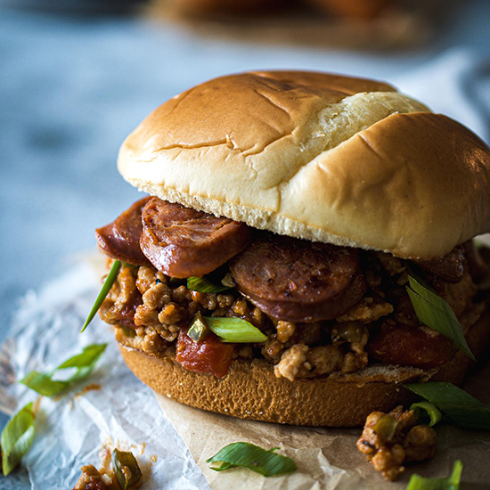 A sloppy joe sandwich with slices of sausage on a brioche roll sitting on parchment paper.