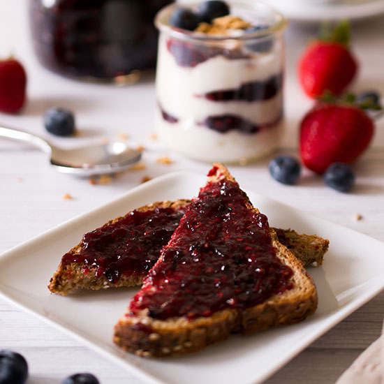 Toast covered in homemade jam, with a yogurt parfait and fresh berries in the background.