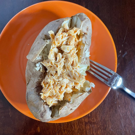 Shredded buffalo chicken on a baked potato served on an orange plate with a fork