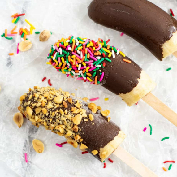 Three frozen bananas on popsicle sticks with nuts, sprinkles and chocolate.