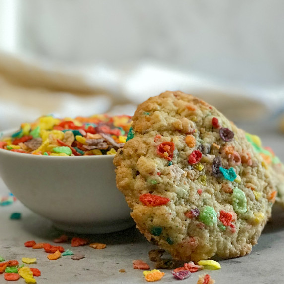 A colorful Fruity Pebbles Cookies leans against a white bowl full of rainbow Fruity Pebbles Cereal. 