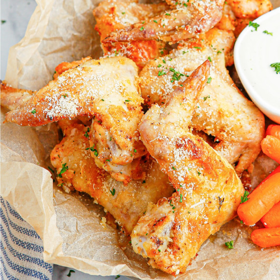 A plate of chicken wings covered in parmesan cheese and carrots with a side of dipping sauce.