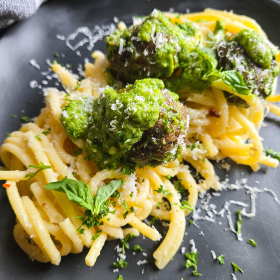 Tender bucatini topped with meatballs tossed in vibrant green pesto on a black plate.