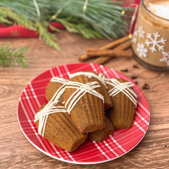 gingerbread madeleines stacked on a red plaid plate with coffee, winter greenery, & cinnamon sticks