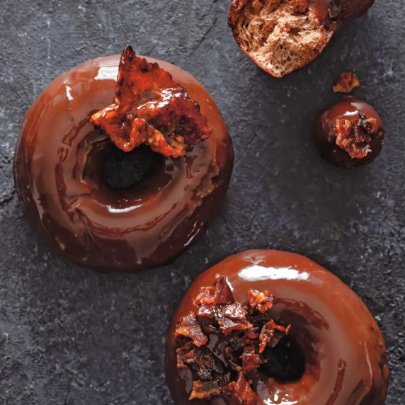 Chocolate-glazed donuts with crispy bacon, one broken to show its soft interior.