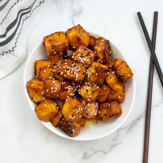 Golden brown Gochujang Tofu cubes garnished with sesame seeds in a white bowl, with chopsticks