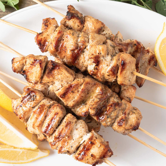 A white platter of Greek chicken kebobs with lemon wedges and fresh oregano.