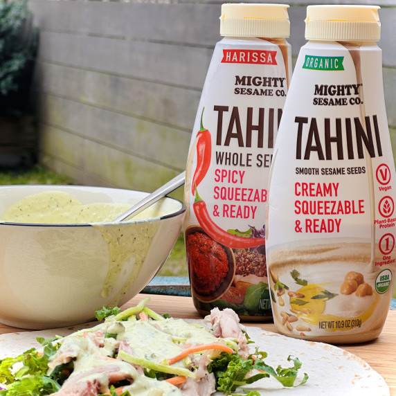 Green tahini sauce in a bowl and drizzled over a salad, next to two bottles of Mighty Sesame tahini
