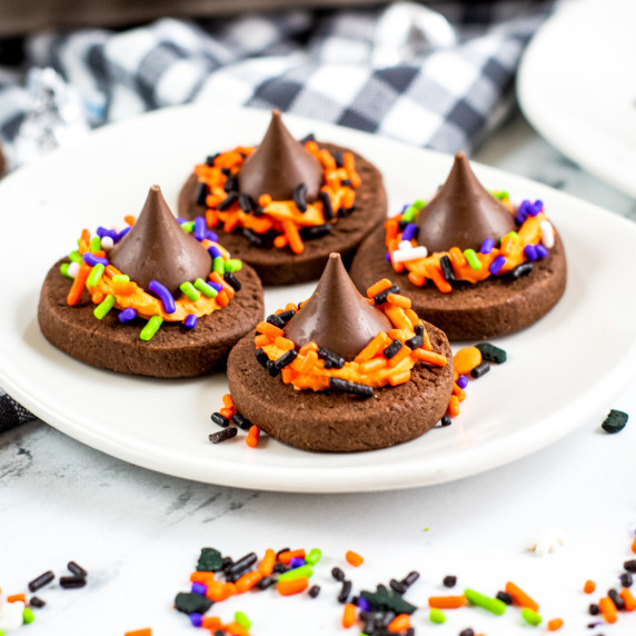 Four chocolate witch hat cookies with Hershey kisses on a white plate with halloween sprinkles