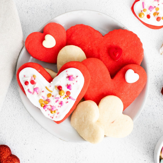 Red and plain heart-shaped cookies on a white plate. Some are decorated with icing and sprinkles.