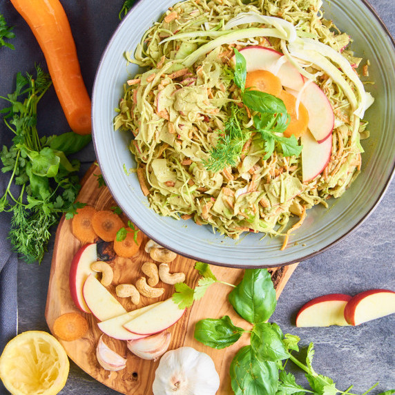 A bowl of Herbed Cashew Aioli Slaw surrounded by apples, herbs and garlic