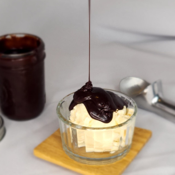 Hot fudge drizzled on vanilla ice cream with a jar of sauce and an ice cream scoop in the background