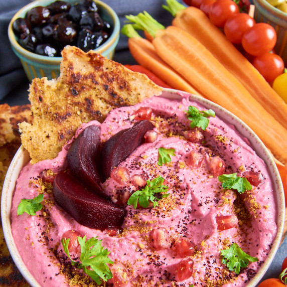 A bowl of creamy and vibrantly pink Beetroot Hummus surrounded by crudites. Plain hummus in the back