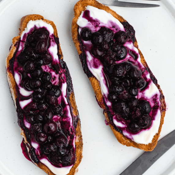 Two toasted bread topped with yogurt, warm blueberries, and honey drizzle on a white plate