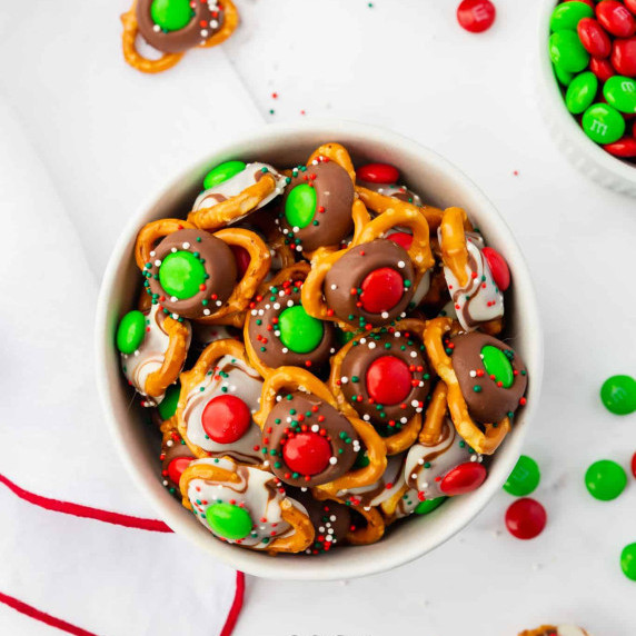 Pretzels topped with Hershey's chocolate kiss, green or red M&M's, and sprinkles in a white bowl