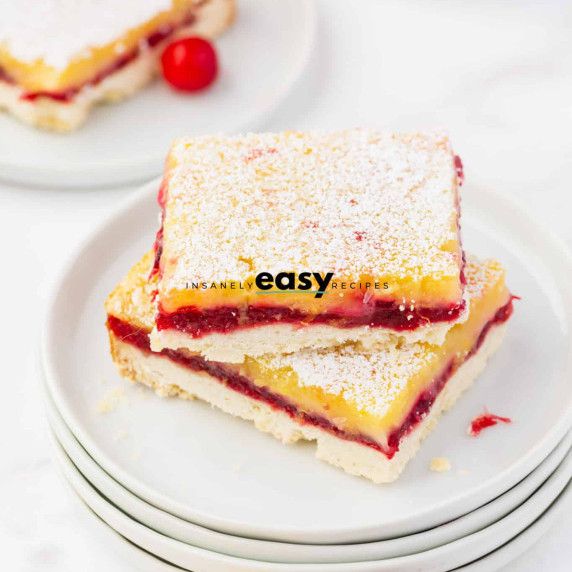 Two lemon bars with a layer of cranberry jam in the middle on top of stacked white plates