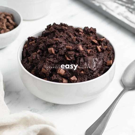 Edible brownie batter with chocolate chunks in a white bowl, a spoon and dish towel next to it 