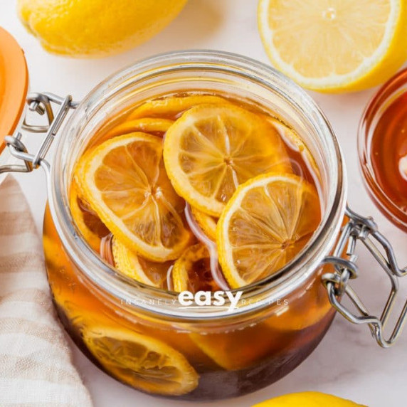 Glass jar of honeyed lemon slices, with fresh lemon fruits and a bowl of honey on the side