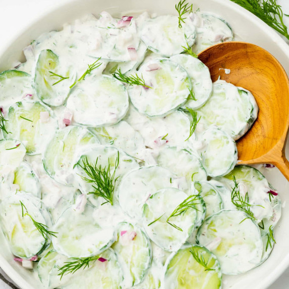 Cucumber salad with sour cream topped with additional dill in a white bowl with a wooden spoon