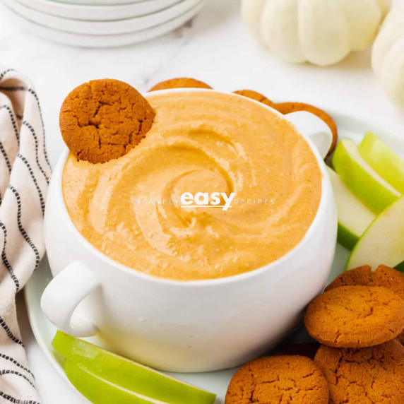 Pumpkin cream cheese dip in a white bowl served with ginger cookies and sliced apples