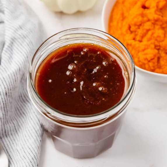 Pumpkin spice syrup in a glass container with a bowl of pumpkin puree behind and a dish towel beside