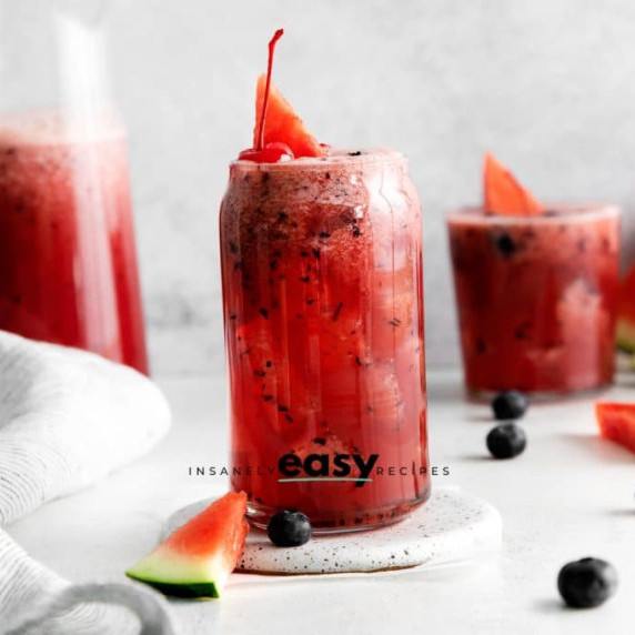 A tall glass, a regular glass, and a pitcher of red cocktail with watermelon, cherres, & blueberries