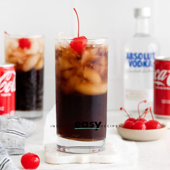 Tall glass of coke and vodka cocktail with ice and cherry on top, Coke cans and vodka bottle behind 