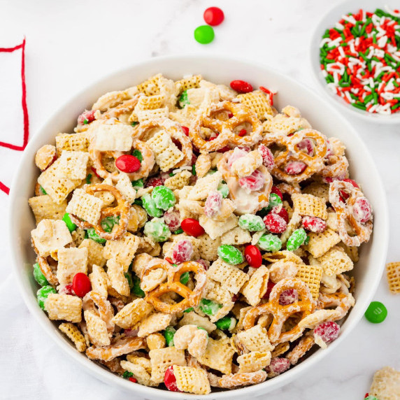 Chhristmas-themed Chex mix in a white bowl with a smaller bowl of sprinkles beside on a countertop