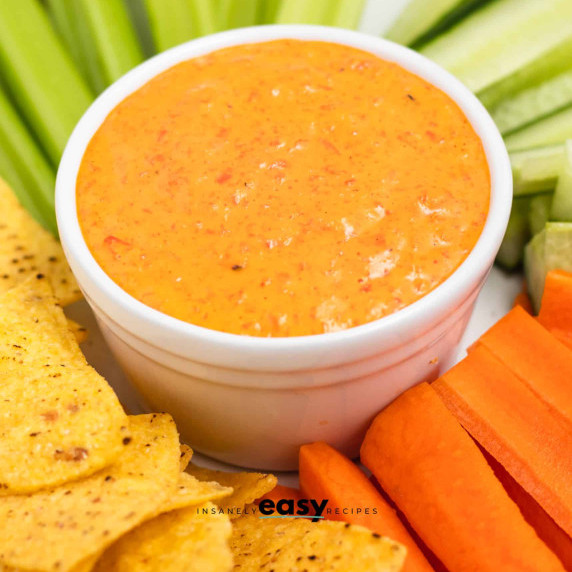 Red pepper crema in a white bowl surrounded by chopped vegetables and chips