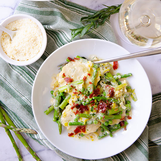 Bowl of ravioli and asparagus next to grated parmesan and glass of white wine.