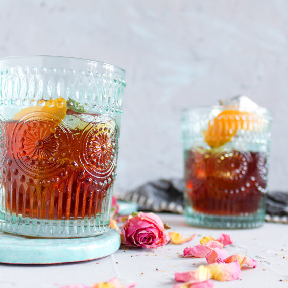 brown bourbon in blue glasses with red and yellow rose petals sprinkled around on grey background