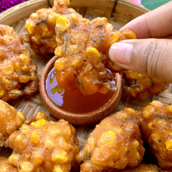 Hand dipping Thai corn fritters into sweet chili sauce, with more corn fritters around it.
