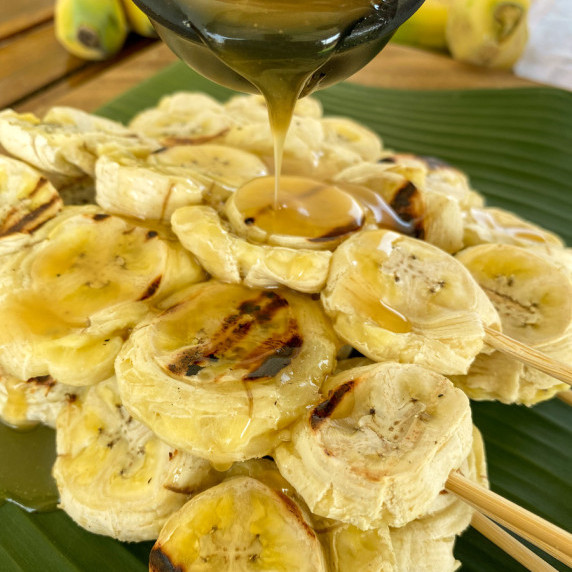 Grilled bananas on a skewer with a black sauce cup over it that's pouring caramel sauce on top.