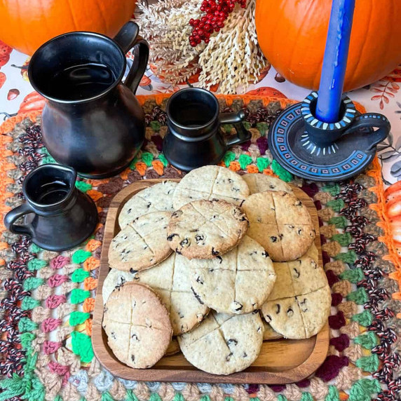 A pile of soul cakes on a wooden platter with candle to side and pewter jug, and pumpkins.