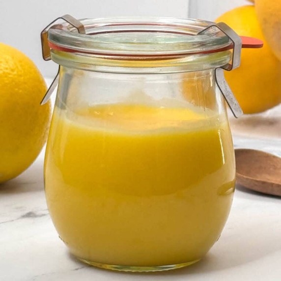 Side view of a fancy glass jar of homemade lemon curd with lemons and a spoon in the background 