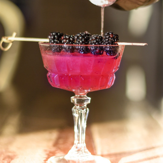 Gold cocktail shaker pouring a blackberry martini into a glass, with blackberry garnish.