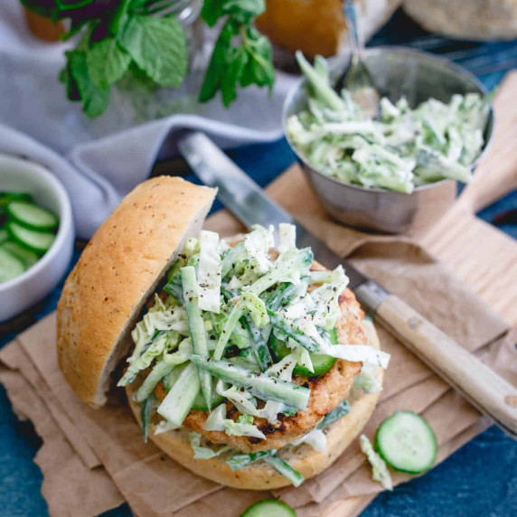 A grilled chicken burger on a bun is topped with a creamy coleslaw and cucumber slices