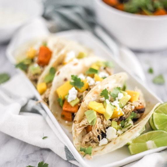 Chicken tacos on a rectangle plate topped with mango, avocado, cilantro and cotija cheese on counter