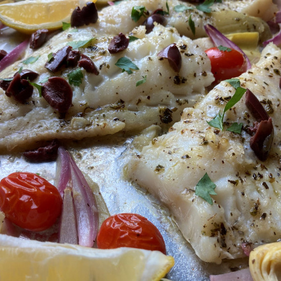 Baked cod on a sheet pan with roasted tomatoes, red onion, black olives, and lemon wedges.