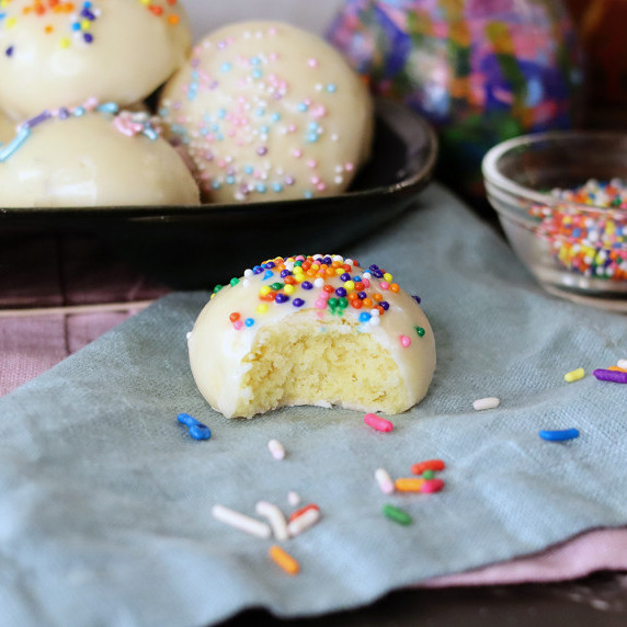 Italian sprinkle cookie with a bite missing on a blue cloth napkin, more cookies on a plate behind.