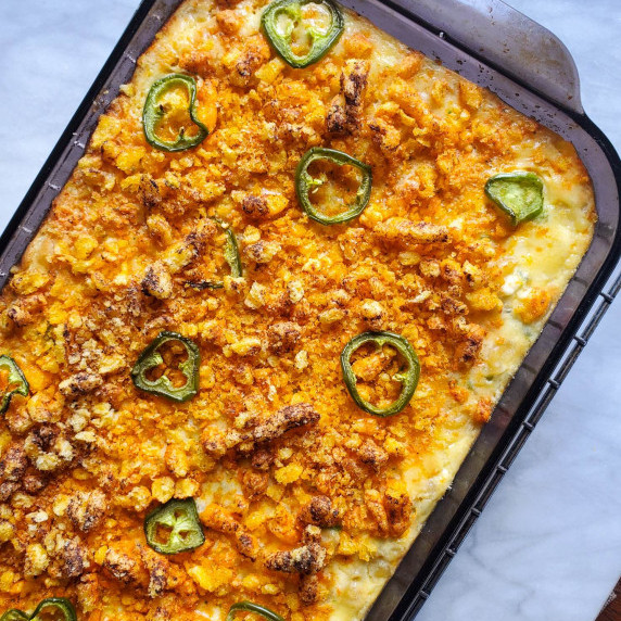 A cheesy, cheetos topped mac and cheese with sliced jalapenos against white marble.