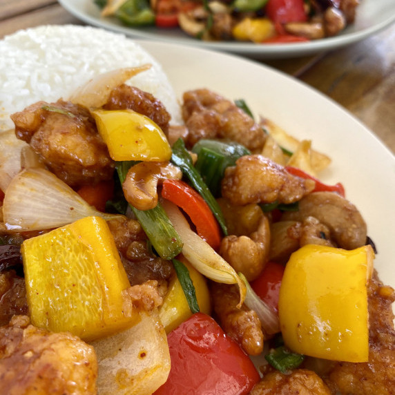 Stir-fried chicken with cashews and bell peppers on a white dish with white rice next to it.