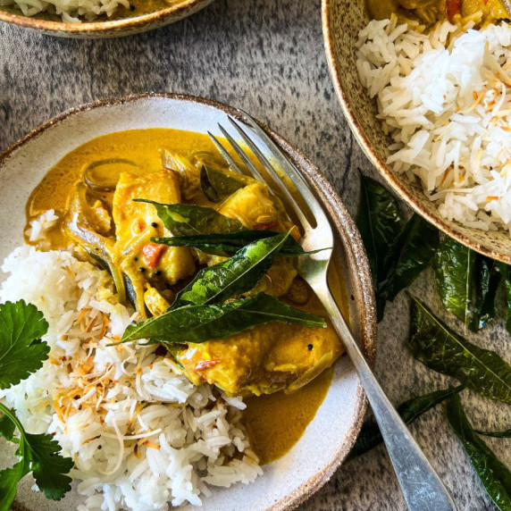 Indian Fish Curry on a plate with rice, fork and green garnish. 2 smaller bowls to the side